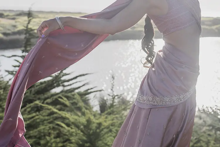 Cuerpo de una mujer vestida con un saree. no se le ve el rosto. Se encuentra en un espacio verde con el sol de fondo.